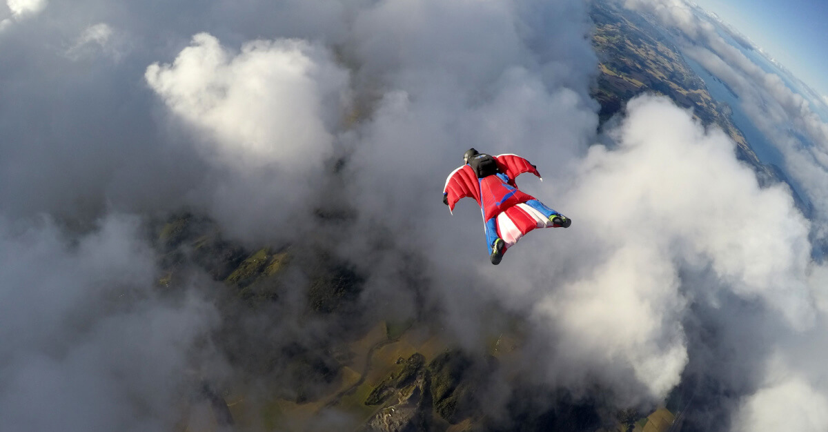 A person, suspended among clouds by a red wingsuit. Exterior, day.